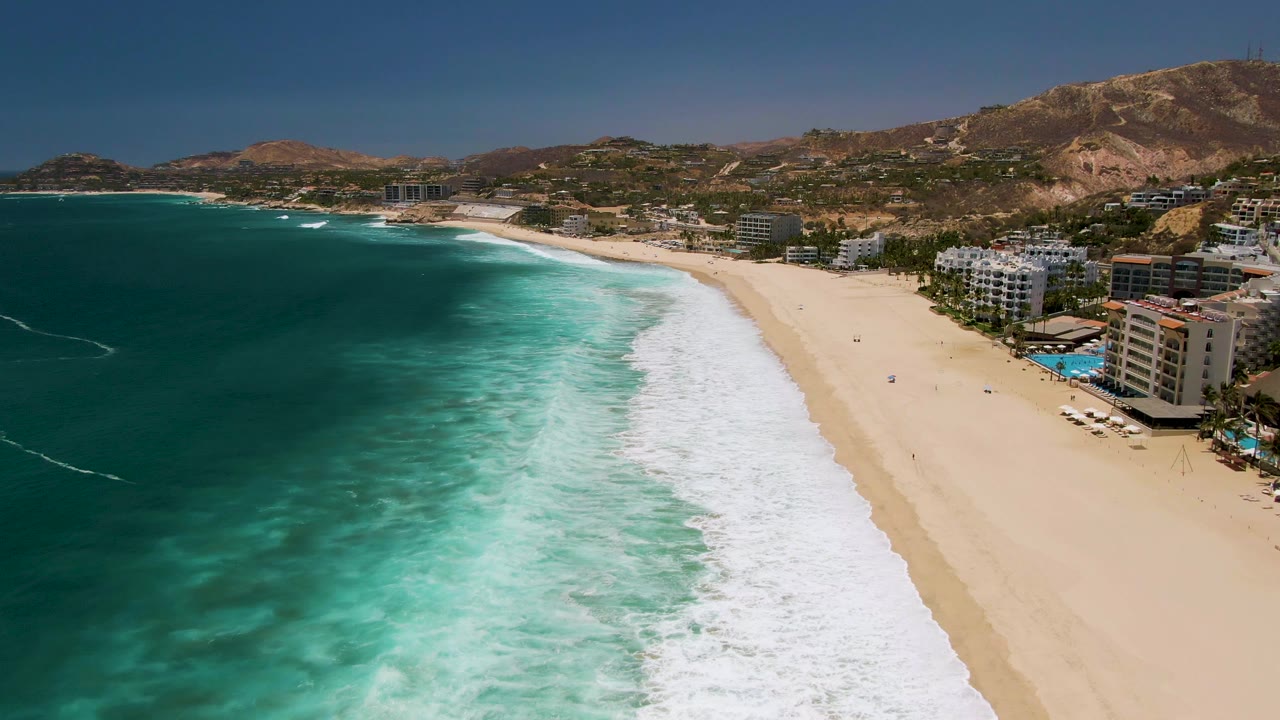 Wide drone view of surf spreading across sand near beachside buildings.