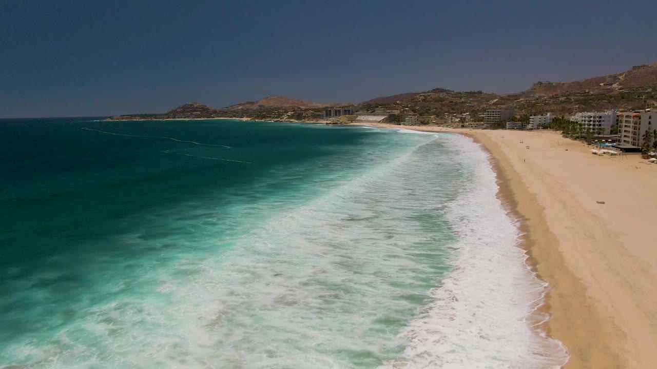 Aerial frame of Costa Azul Beach in Los Cabos with turquoise water, white foam, a sandy shoreline, distant coastal buildings, dry hills, and no visible people.