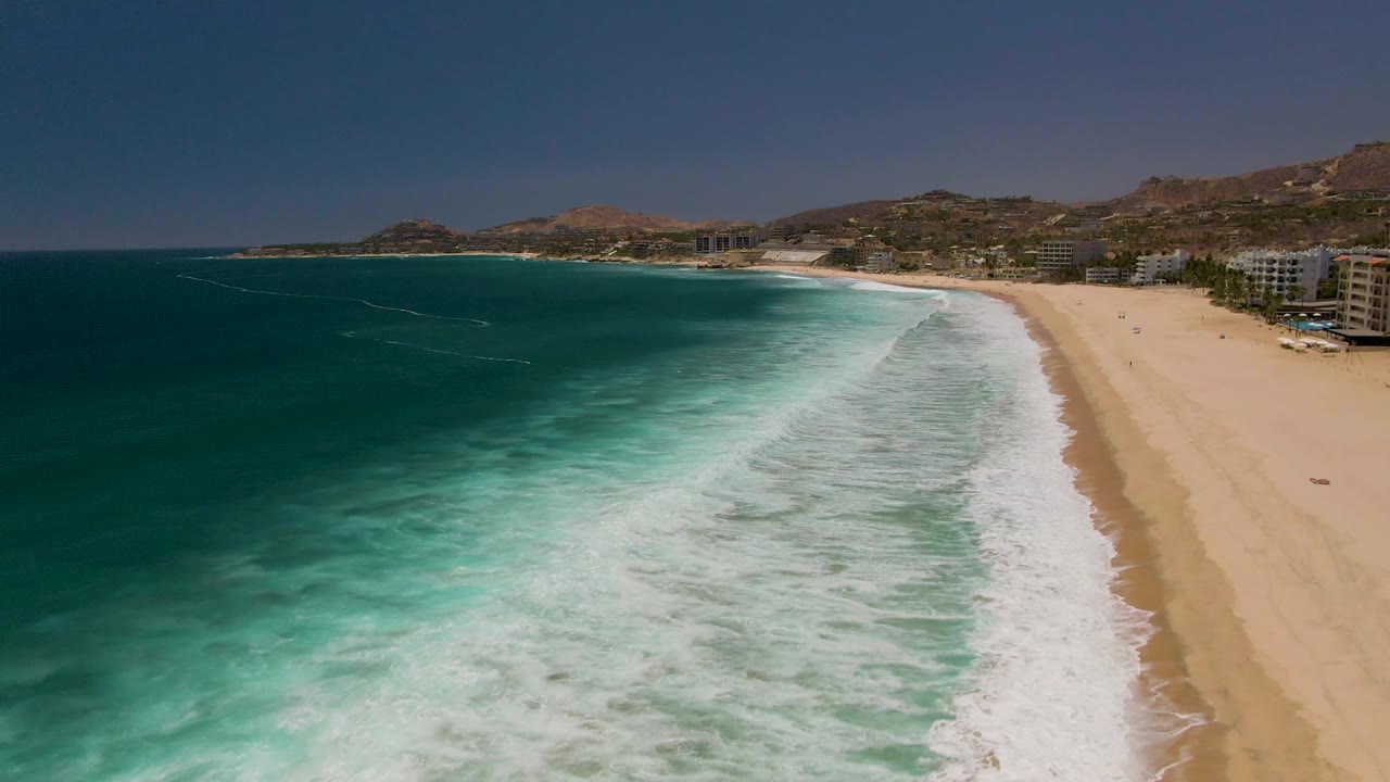 Aerial view over Costa Azul Beach in Los Cabos as white surf spreads across shallow turquoise water beside a broad sandy beach with no visible people.