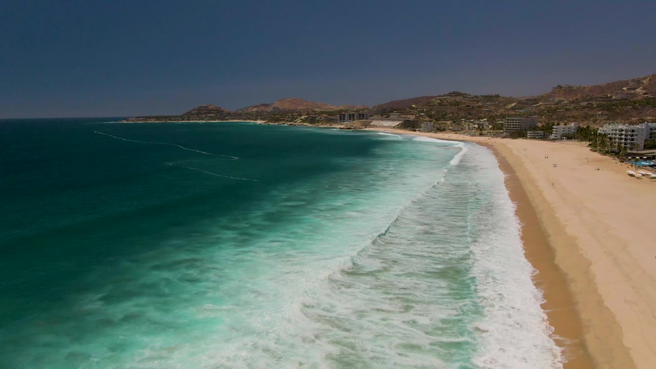 Costa Azul Beach in Los Cabos seen from above with layered surf bands, a tan shoreline, coastal buildings to the right, low hills in the distance, and no visible people.