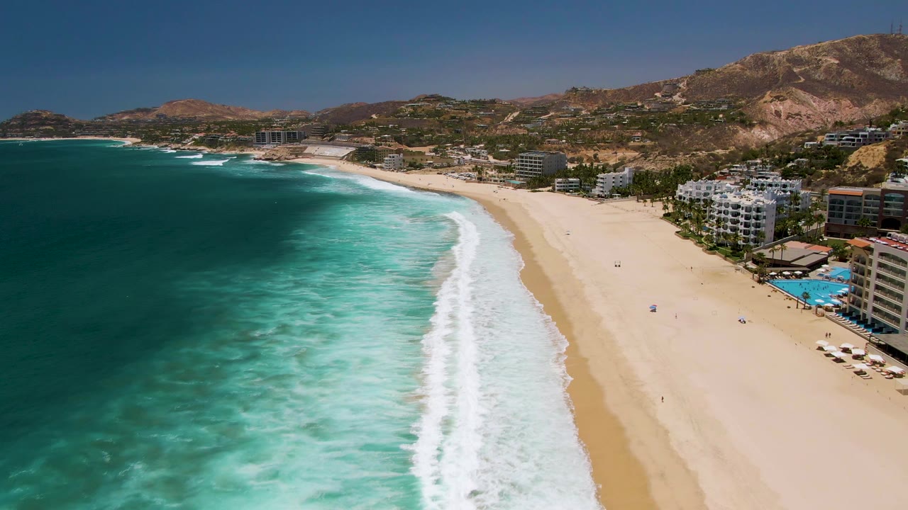 Turquoise surf lines a broad sandy beach with hotels and dry hills beyond.