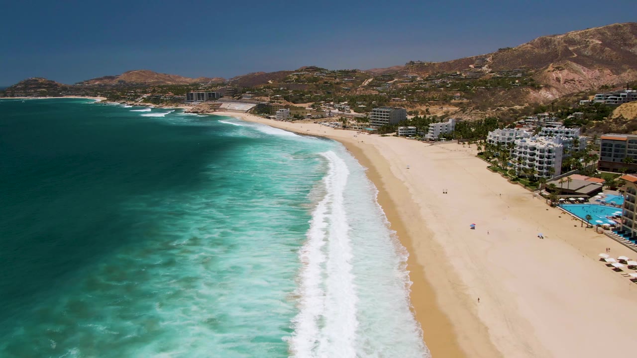 Aerial view over Costa Azul Beach with foamy surf running toward the lower frame, open sand on the right, clustered coastal buildings near the shoreline, and tiny people visible on the beach.