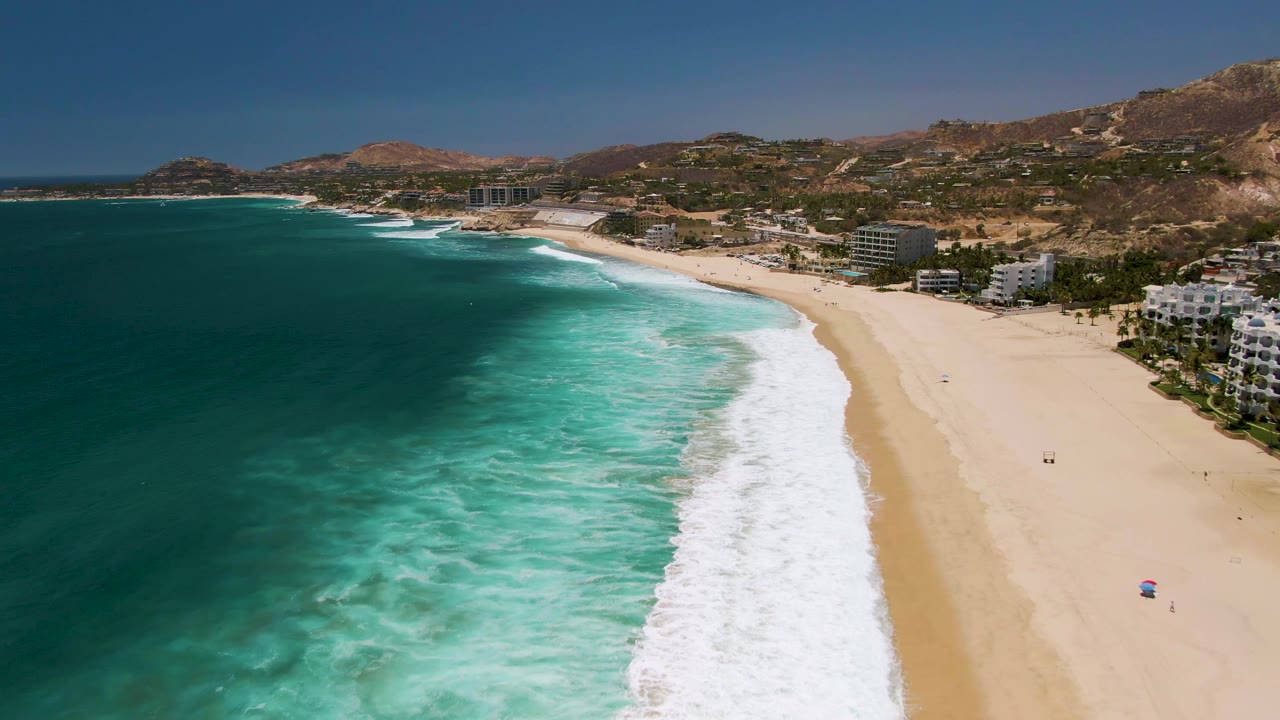 Aerial view over Costa Azul Beach in Los Cabos, with deep blue water at left, turquoise surf rolling onto pale sand, coastal buildings on the right, hills in the background, and small people visible on the beach.