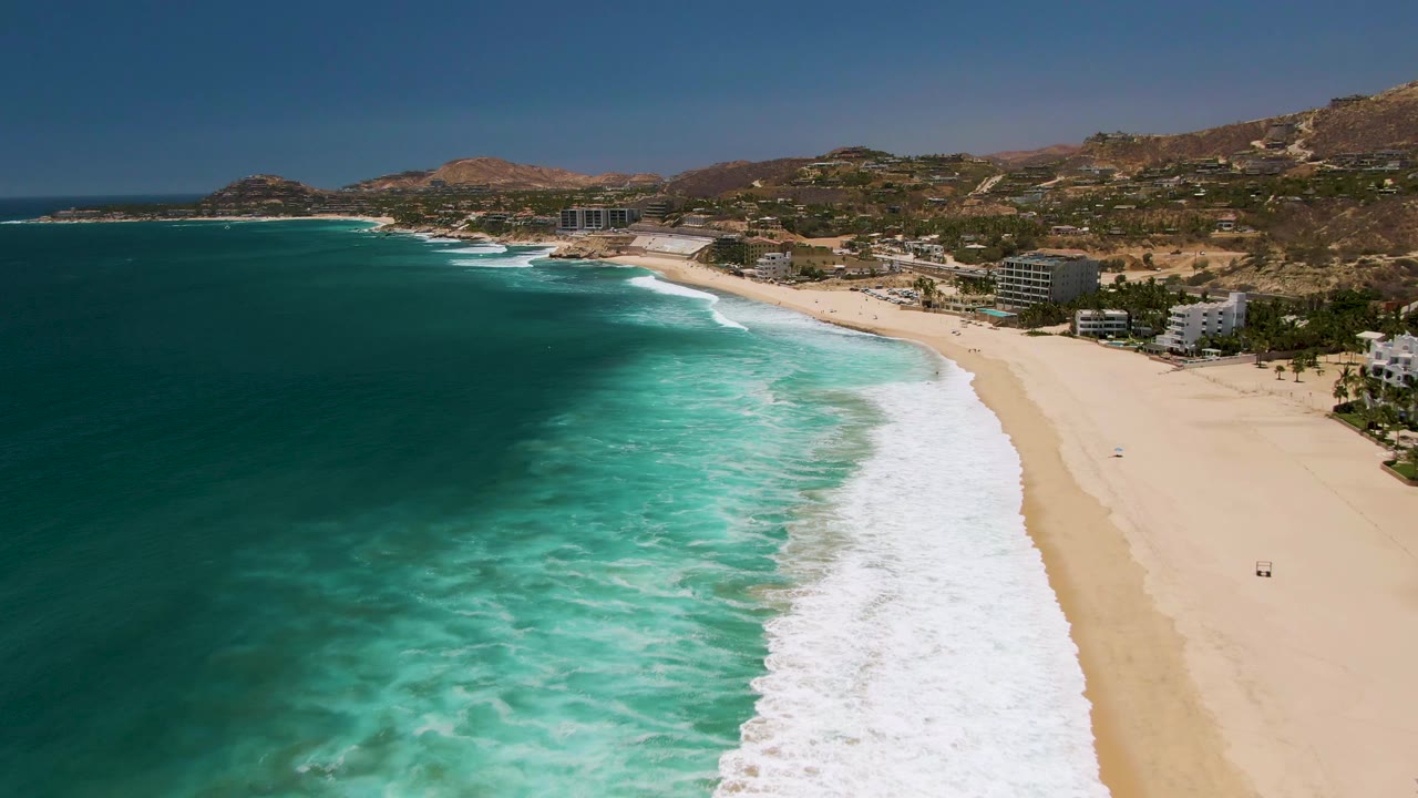 Wide aerial frame at Costa Azul Beach in Los Cabos with foamy surf stretching across the center, open sand to the right, darker blue water to the left, coastal buildings near the shoreline, and hills under clear sky.