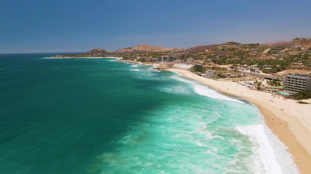 At 0:00, Costa Azul Beach Los Cabos is seen from above with turquoise water in the foreground, white surf at the shoreline, small beachgoers on the sand, and dry hills behind coastal buildings.
