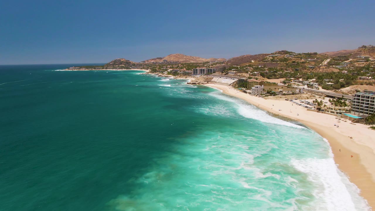 Wide aerial coastline view with deep blue ocean, pale beach, breaking waves, buildings, and hills.