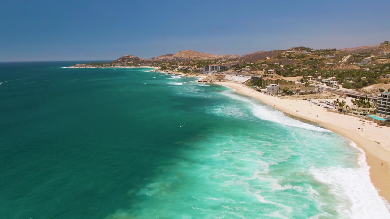At 0:02, foamy turquoise water occupies more of the lower frame while the sandy shore, small figures, road activity, coastal buildings, and arid hills remain visible along Costa Azul Beach Los Cabos.