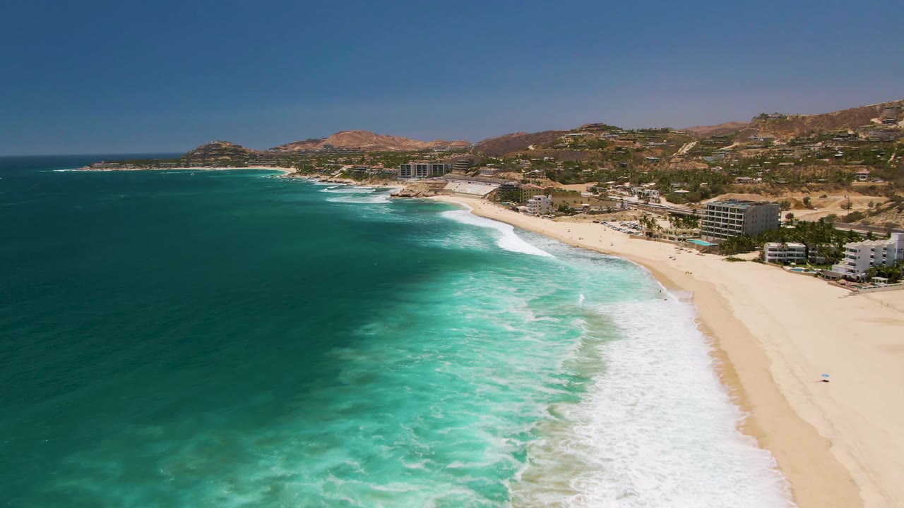 Opening aerial frame over Costa Azul Beach in Los Cabos, with deep blue ocean at left, white surf on pale sand, coastal buildings near the shore, dry hills in the background, and no visible people.