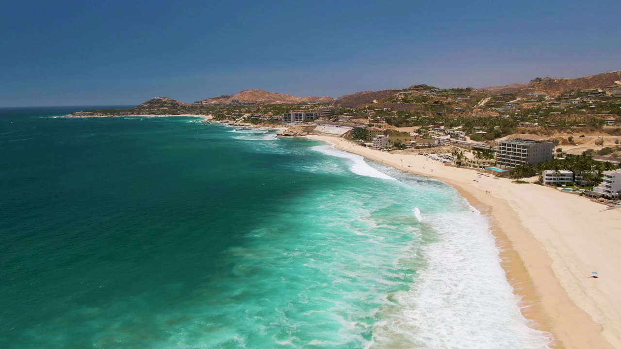 Aerial frame at Costa Azul Beach showing a broader band of white foam rolling shoreward, turquoise water in the foreground, sand to the right, coastal buildings along the beach, and no visible people.