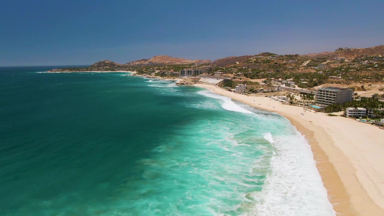 Costa Azul Beach appears from a high aerial angle with clear green-blue water across the foreground, a long white wave line beside the sand, beachfront buildings, hillside terrain, and no visible people.