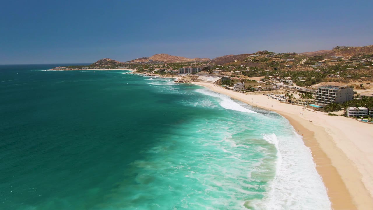 Wide aerial coastline with dark blue ocean, turquoise shallows, foamy waves, sandy beach, and hillside buildings.