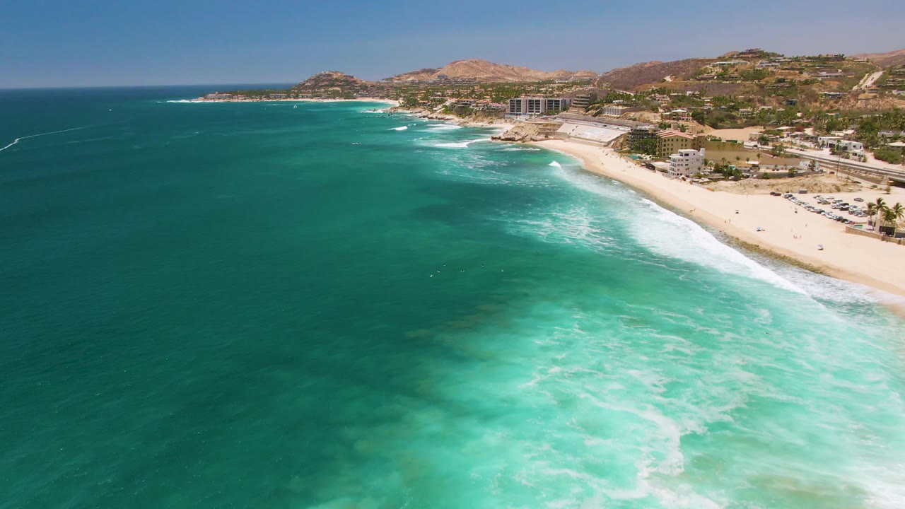 Wide aerial frame over Costa Azul Beach in Los Cabos, with turquoise water in the foreground, white surf reaching the sand, hillside buildings, and tiny beachgoers along the shore.
