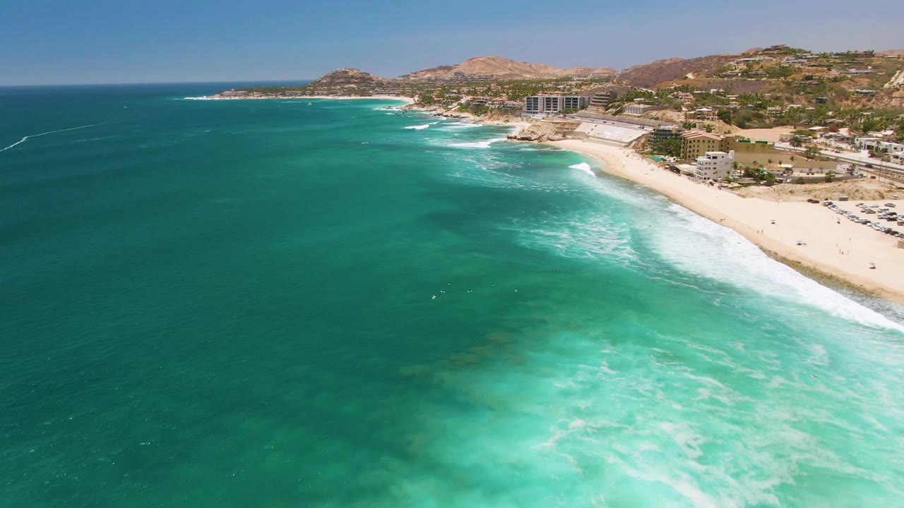 The coastline at Costa Azul Beach in Los Cabos fills the frame, with darker offshore water on the left, bright turquoise shallows below, and a narrow sandy beach beside hillside buildings.