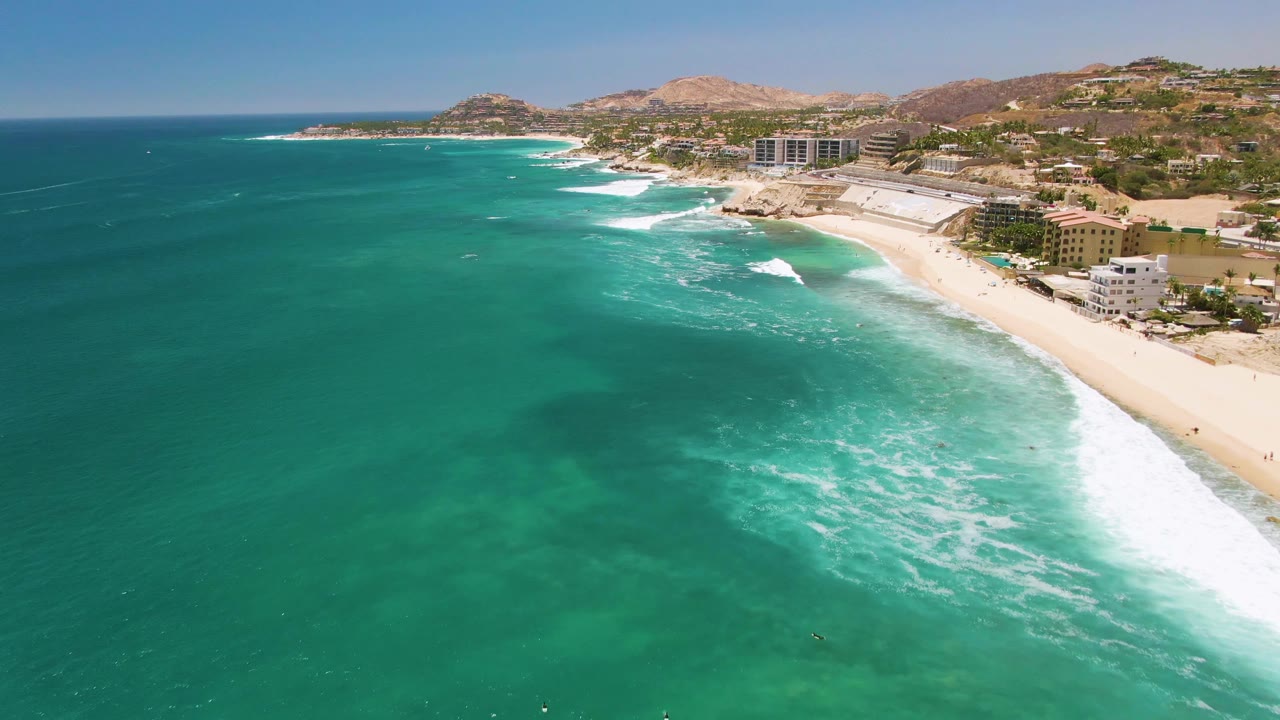 Aerial view over Costa Azul Beach in Los Cabos, with turquoise water in the foreground, white surf nearing the sand, coastal buildings along the right edge, and no visible people.