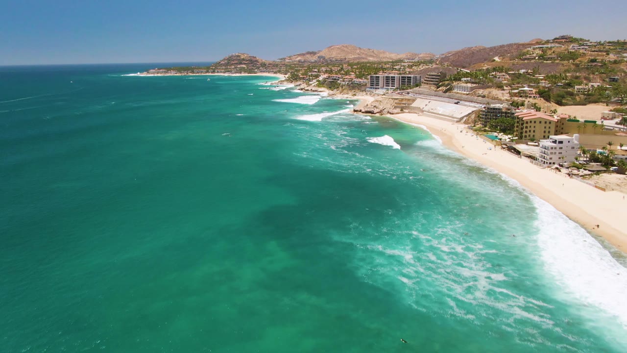 Turquoise water and white waves line a sandy beach beside low coastal buildings.