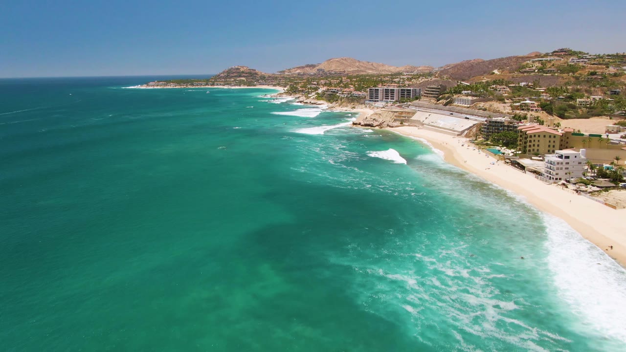 Aerial view of Costa Azul Beach in Los Cabos with clear green-blue water dominating the frame, foamy waves breaking diagonally, a pale beach at right, coastal buildings, dry hills, and no visible people.