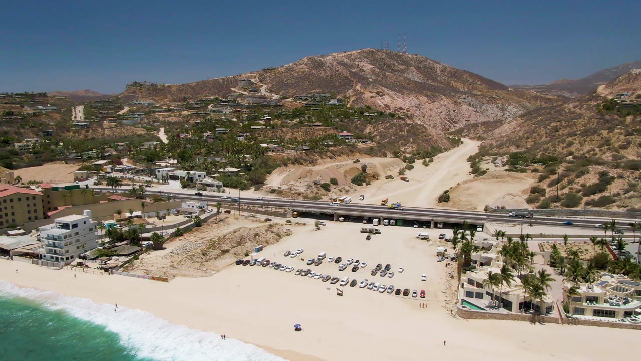 Aerial frame of Costa Azul Beach in Los Cabos with turquoise surf at the lower left, a sandy parking lot full of cars, a raised highway, hillside buildings, dry hills, and no visible people.