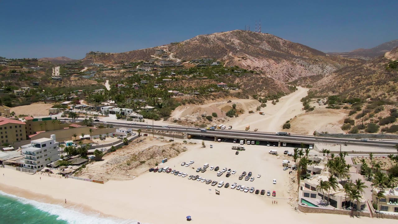 Aerial view at Costa Azul Beach in Los Cabos with the beach parking lot centered, the highway running across the frame, palms and low structures near the sand, dry hills behind, and no visible people.