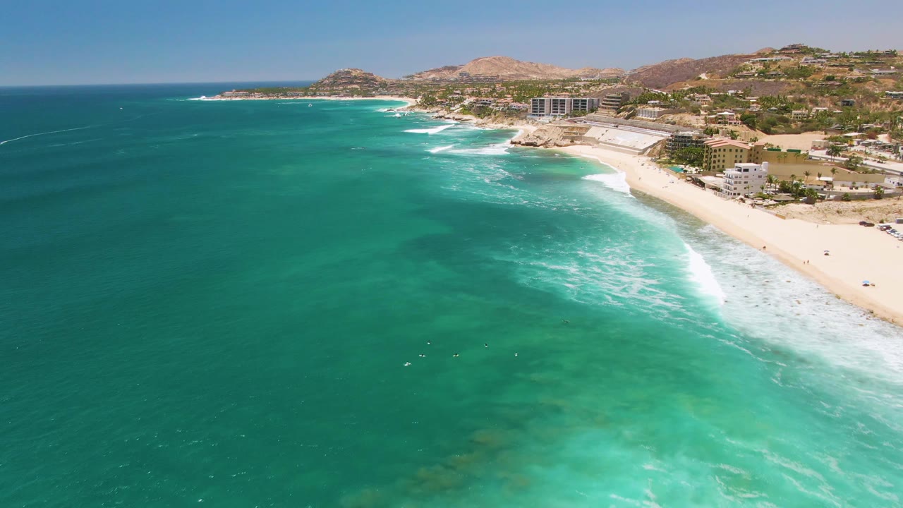 Aerial frame over Costa Azul Beach in Los Cabos, with turquoise water in the foreground, white surf along the sand, hillside buildings in the background, and no visible people.