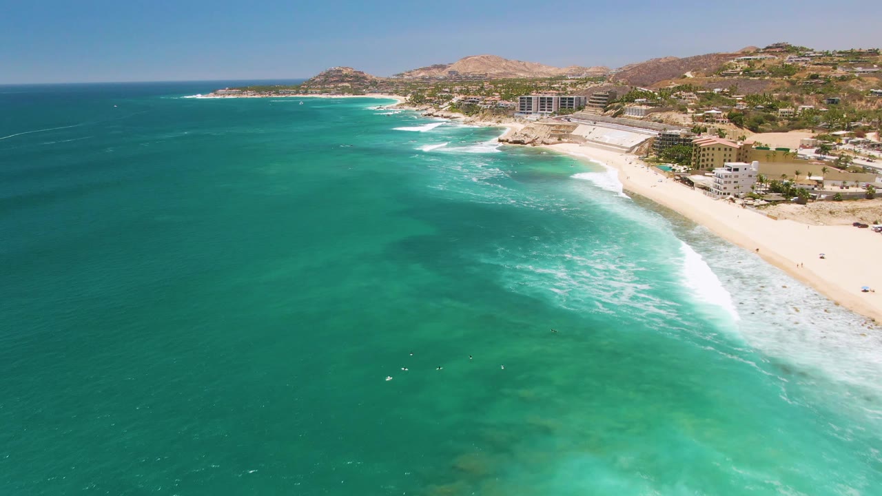 Aerial view at Costa Azul Beach with the surf band slightly farther along the shoreline, clear green-blue water below, coastal buildings beyond the sand, and no visible people.
