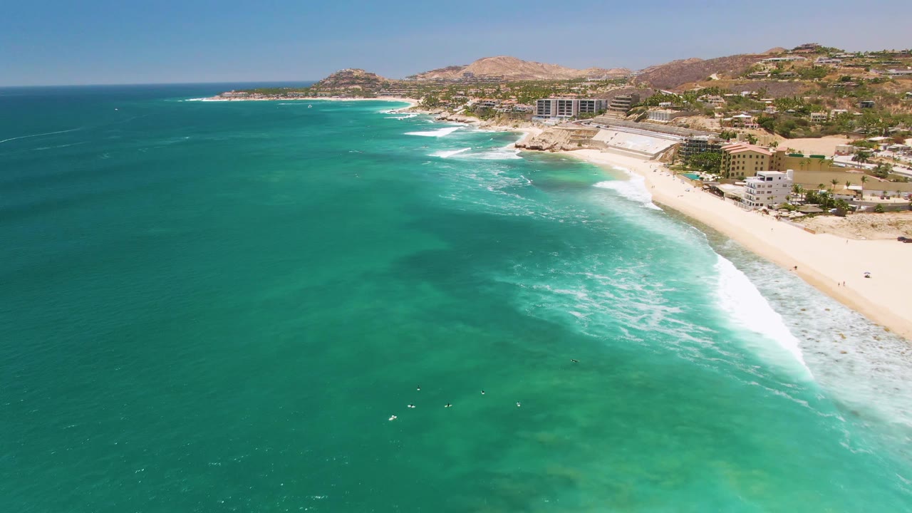 Costa Azul Beach aerial frame showing a larger white breaker near the sand, broad turquoise water in the foreground, hillside development behind the beach, and no visible people.