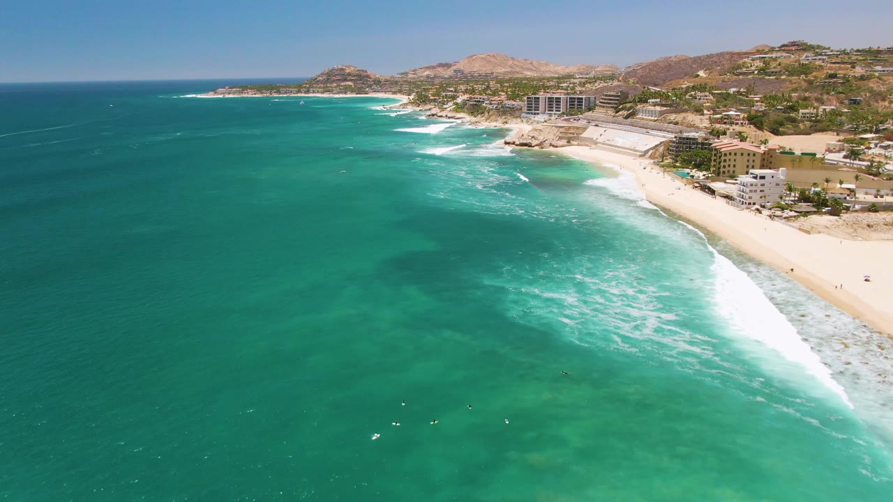 Clear ocean water, white surf, sandy beach, and coastal buildings seen from above.