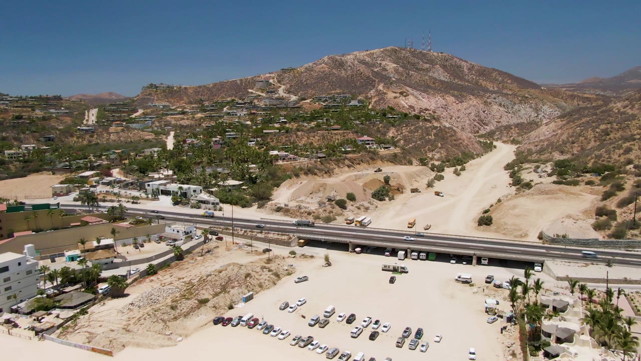 Aerial frame at Costa Azul Beach Los Cabos showing highway traffic, a sandy parking lot with cars, dry hills, scattered buildings, and no visible people.