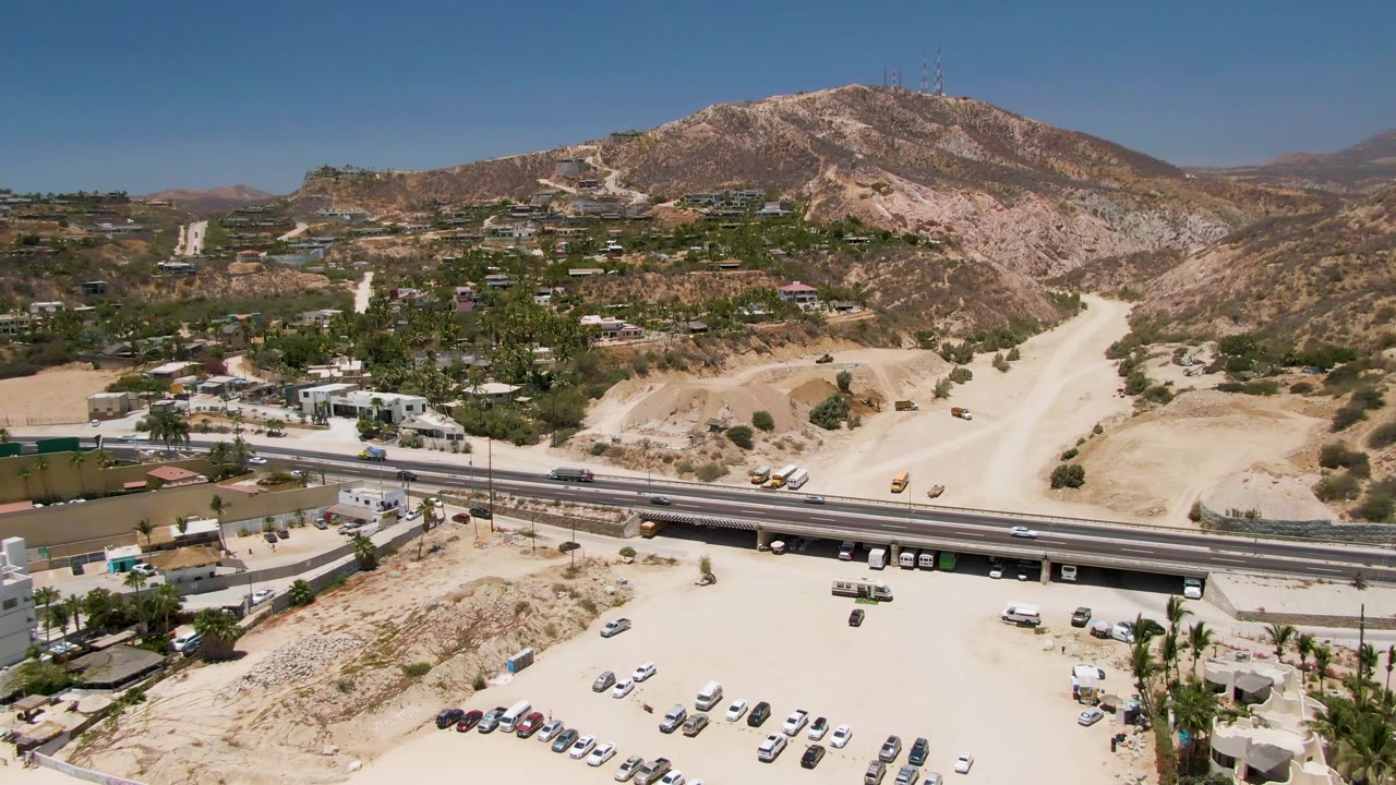 Drone view over Costa Azul Beach Los Cabos with a divided road, passing vehicles, hillside homes and structures, exposed dirt slopes, and no visible people.