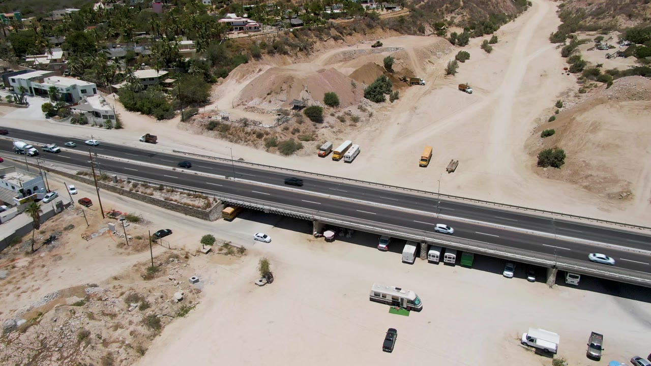 Aerial frame from Costa Azul Beach Los Cabos showing a multi-lane highway over a dirt service area with parked trucks, scattered trees, and no visible people.
