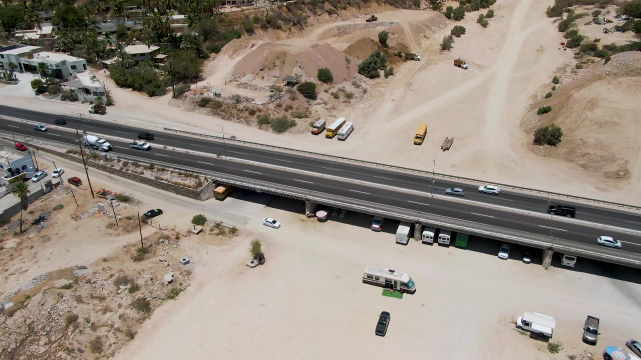Aerial frame from Costa Azul Beach Los Cabos showing the highway centered across dry terrain, with trucks parked under the overpass and no visible people.