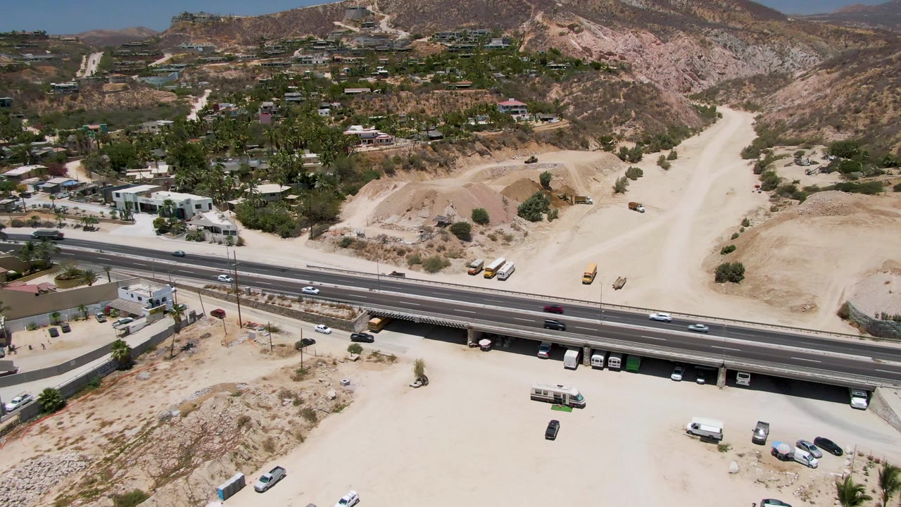 Aerial frame from Costa Azul Beach Los Cabos showing highway traffic beside pale dirt lots, construction equipment, scattered buildings, dry hills, and no visible people.