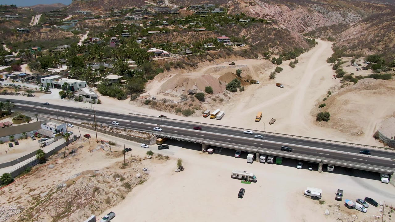 Cars and trucks move along a highway next to dirt lots and hillside buildings.