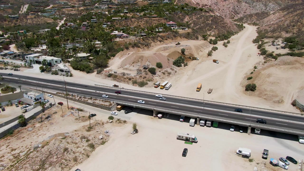 Costa Azul Beach Los Cabos aerial frame with the highway crossing the lower scene, yellow construction equipment on pale dirt, scattered buildings, dry hills, and no visible people.