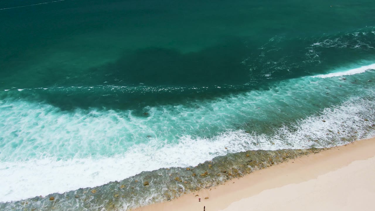 Overhead frame at Costa Azul Beach in Los Cabos shows dark teal ocean, white foam, rocky shallows, pale sand, and tiny people near the lower edge.