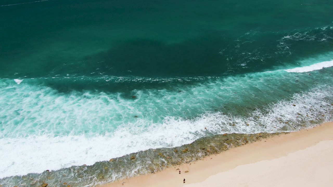 Turquoise surf spreads across rocks beside pale sand