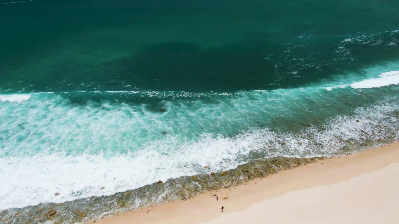 At 0:02, foamy surf runs diagonally across the middle of the Costa Azul Beach frame, separating deeper blue-green water from the sandy shore and tiny figures.