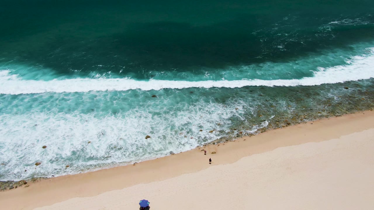 Overhead view of Costa Azul Beach in Los Cabos with white surf rolling toward pale sand, two small people near the waterline, and a blue umbrella at the lower edge.