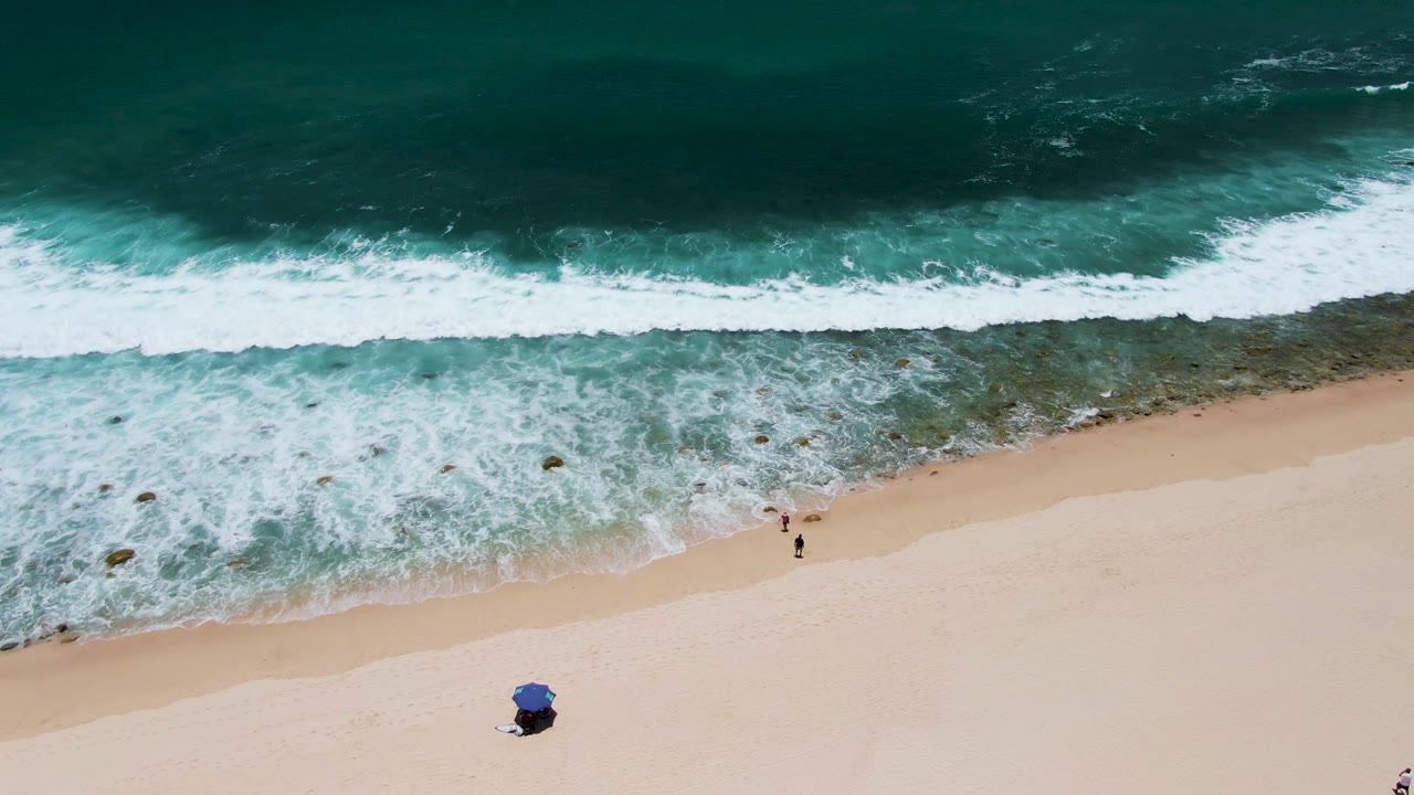 At Costa Azul Beach, a wide band of white water breaks across the top half of the frame while two small figures walk near the wet sand and a blue umbrella sits lower in frame.