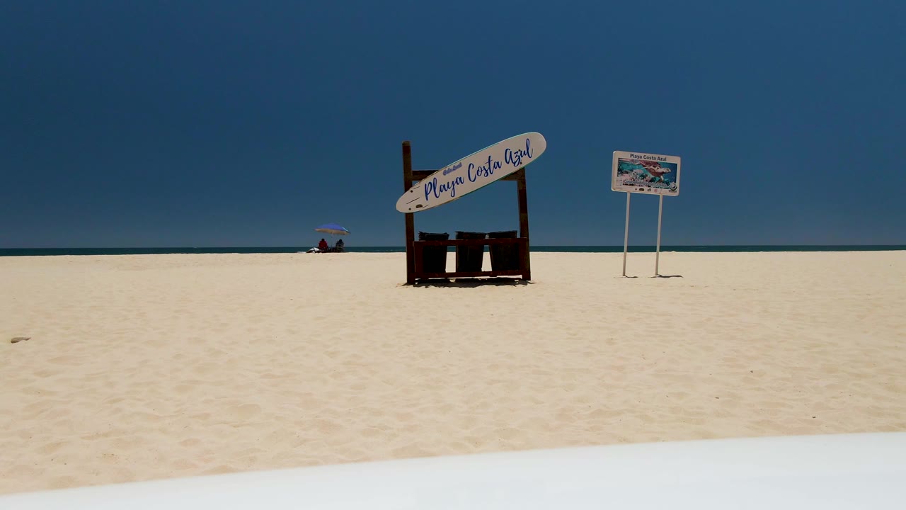 Wide view of Costa Azul Beach in Los Cabos with white sand, a central wooden beach sign, a smaller sign to the right, ocean at the horizon, blue sky, and distant people near an umbrella.