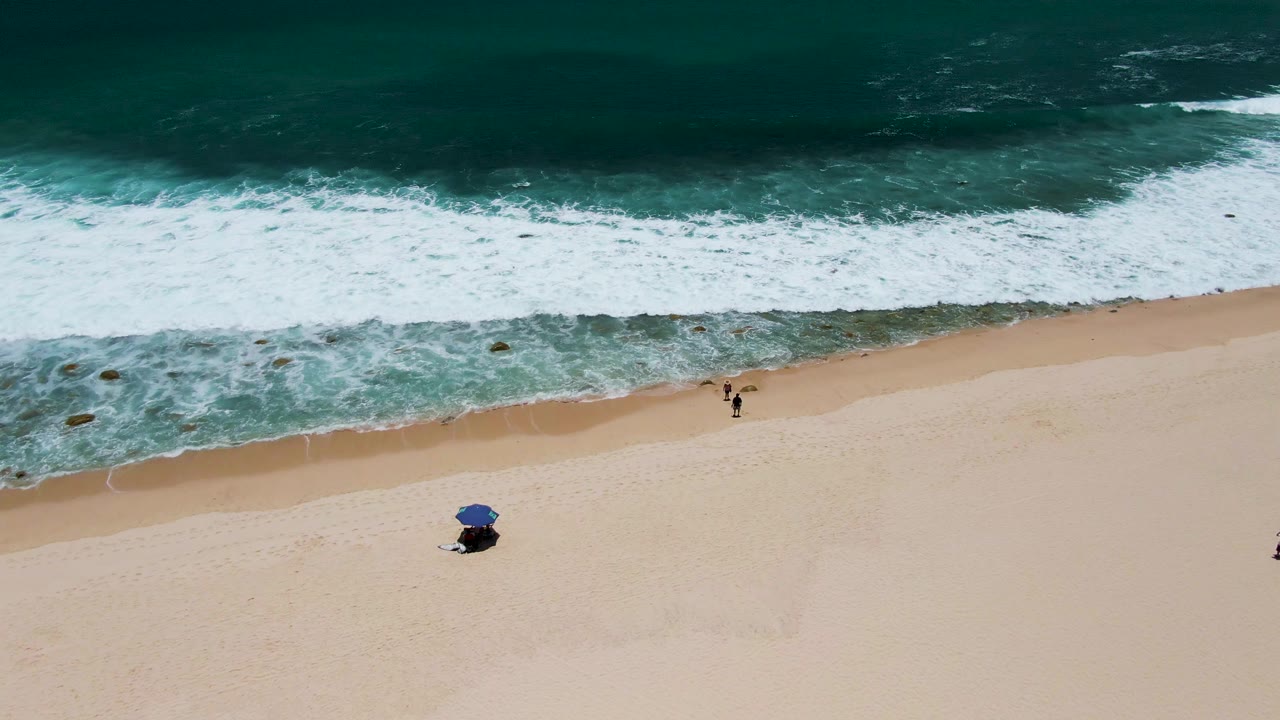 Aerial view of Costa Azul Beach in Los Cabos with dark teal water, white surf, pale sand, a blue umbrella, and small people near the waterline.