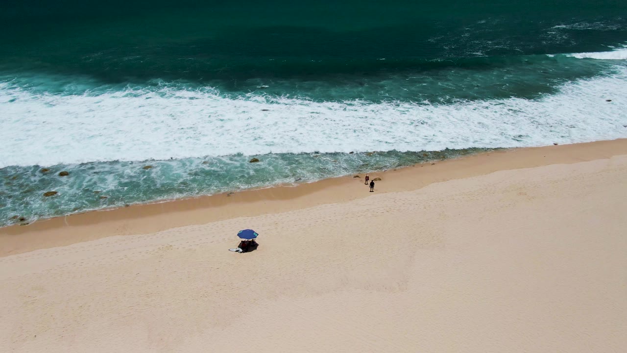Drone view of surf washing onto sand with a blue umbrella and distant beachgoers.