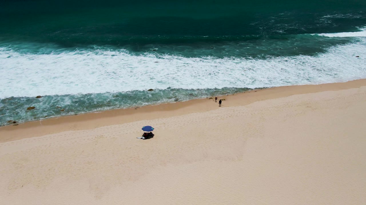 A high aerial frame over Costa Azul Beach with white surf across the upper half, broad pale sand below, a blue umbrella, and two small figures near the waterline.