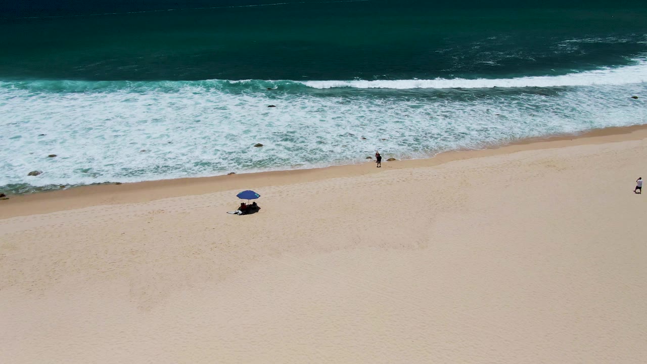 Aerial frame at Costa Azul Beach in Los Cabos showing dark teal ocean, white surf, pale sand, a blue umbrella, and small distant beachgoers near the shoreline.