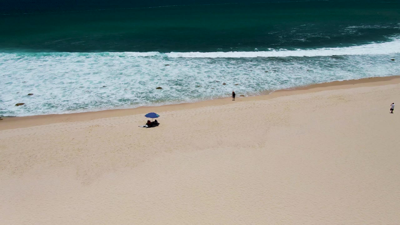 Surf rolls toward a sandy beach with a blue umbrella and distant walkers.