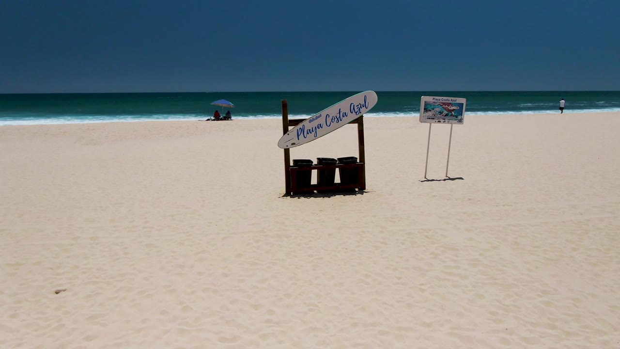 Wide view of Costa Azul Beach in Los Cabos with pale sand, a Playa Costa Azul sign, trash bins, a second sign, ocean horizon, umbrella figures at left, and a small person near the right waterline.