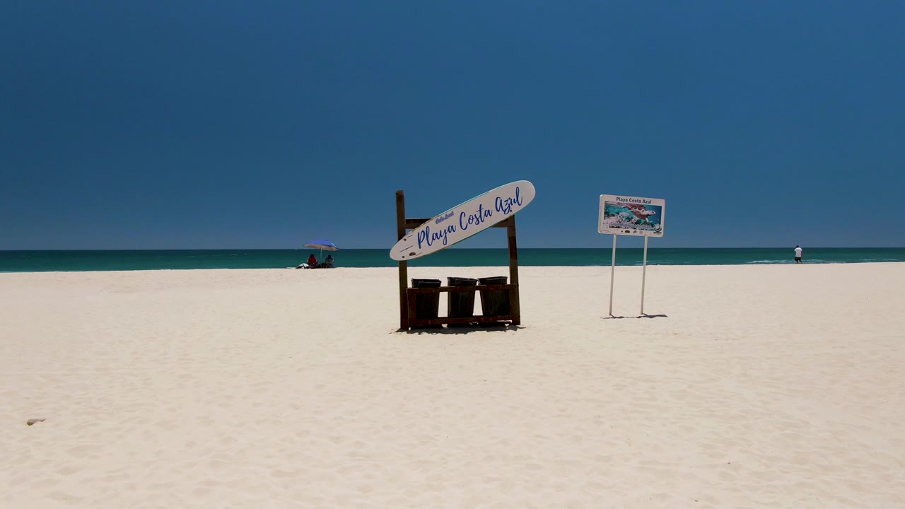 Wide Costa Azul Beach view with the main wooden sign and bins near the right half of frame, a second sign farther right, open white sand in front, turquoise water behind, and distant people near both sides.