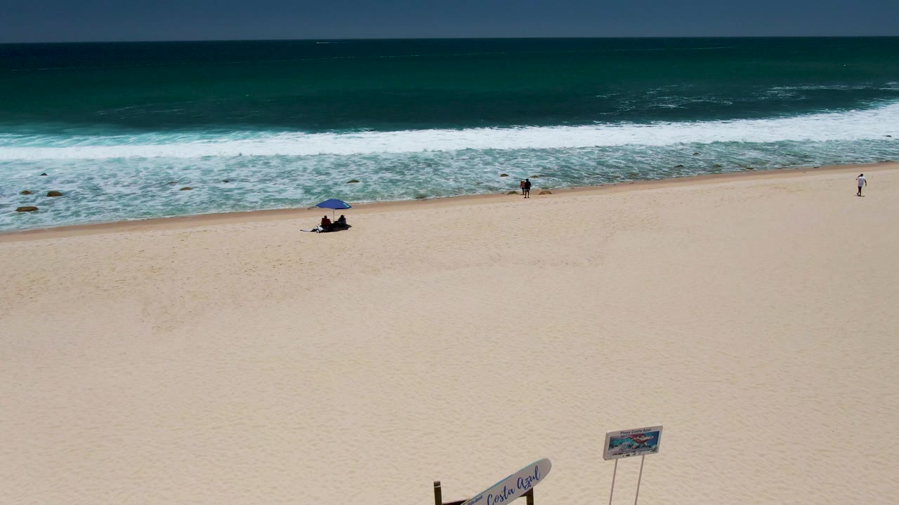 Costa Azul Beach in Los Cabos seen from above, with pale sand in front, blue-green waves across the top of frame, small people near the shoreline, and only the top of a beach sign visible at the lower edge.