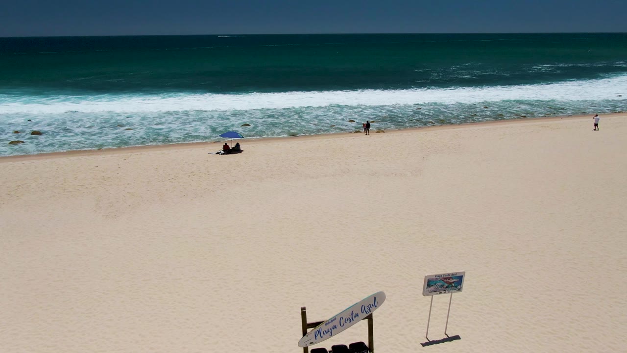 Sandy beach and surf with seated beachgoers, a walker near the water, and a partly visible foreground sign.