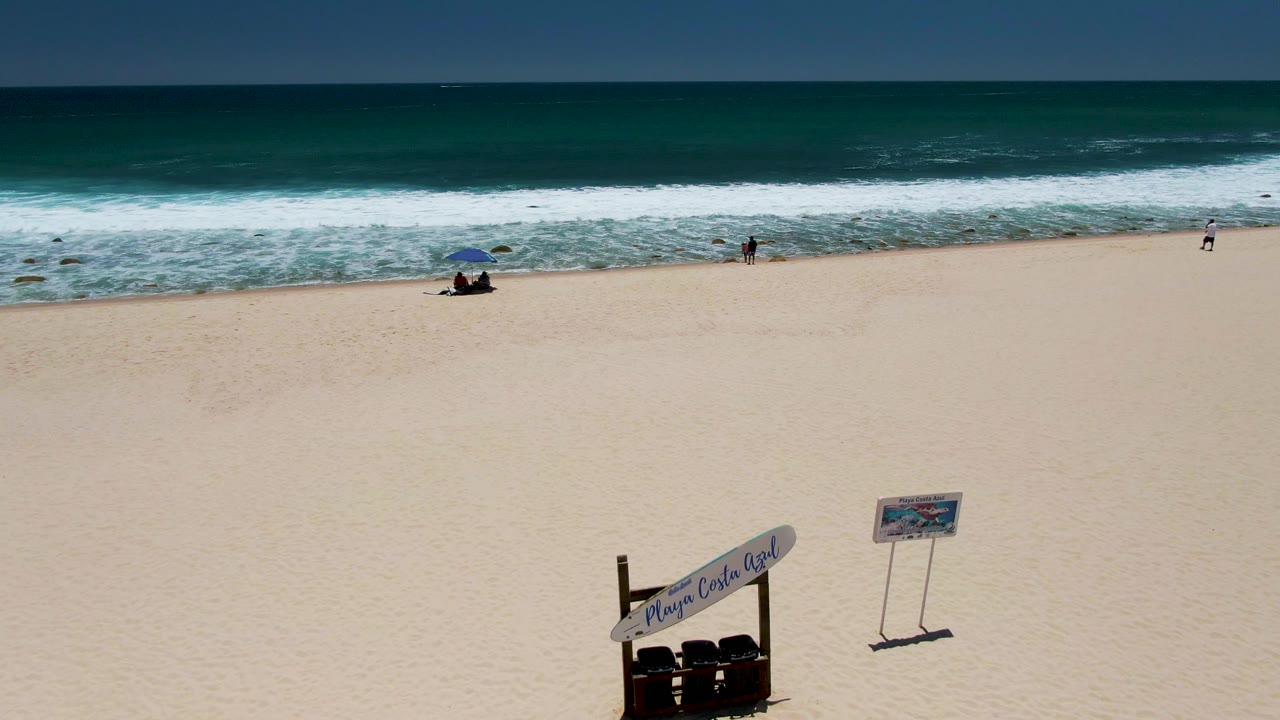 Costa Azul Beach view with broad empty sand, white surf lines, small people spaced near the water, and foreground beach signage becoming easier to read near the lower right.