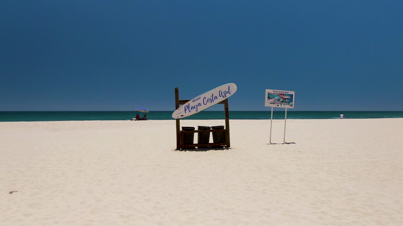 At Costa Azul Beach in Los Cabos, a wide frame shows white sand, a wooden beach sign, a posted sign, turquoise ocean, and a distant umbrella near the shoreline.
